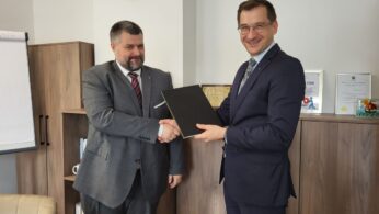 Dwóch mężczyzn w garniturach podaje sobie ręce i uśmiecha się w biurze. Jeden z nich trzyma czarną teczkę. Za nimi znajdują się drewniane szafki, zegar pokazujący 11:10, flipchart i tabliczki z nagrodami. Scena sugeruje formalną umowę lub partnerstwo.