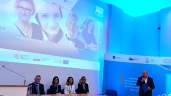 Mężczyzna przemawia na podium w oświetlonej na niebiesko sali konferencyjnej. Cztery osoby siedzą przy stole na scenie. Za nimi na dużym ekranie wyświetlane są cztery zdjęcia i szczegóły wydarzenia w języku polskim, z widocznymi wieloma logo. Atmosfera jest formalna i profesjonalna.
