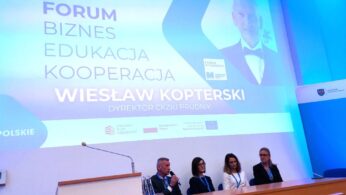 Cztery osoby siedzą przy stole do dyskusji panelowej z mikrofonami przed dużym ekranem. Na ekranie wyświetlany jest polski tekst dotyczący forum na temat biznesu, edukacji i współpracy z udziałem Wiesława Kopterskiego. Otoczenie to nowoczesna, oświetlona na niebiesko sala konferencyjna.