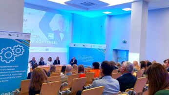 Sala konferencyjna z rzędami siedzących uczestników zwróconych w stronę panelu prelegentów. Wyświetlane są niebieskie banery z białym tekstem i grafiką sprzętu. Duży ekran za prelegentami pokazuje prezentację z portretem mężczyzny i polskim tekstem. Pokój jest jasno oświetlony.