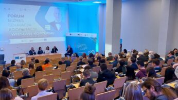 Duża sala konferencyjna z rzędami siedzących uczestników zwróconych w stronę sceny, na której pięciu prelegentów siedzi przy stole. Duży ekran wyświetla tytuł forum w języku polskim. Po bokach widoczne są banery i plakaty z logo wydarzenia.