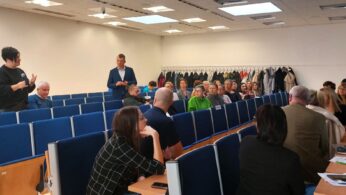 Grupa ludzi siedzi w niebieskich fotelach w sali wykładowej, naprzeciwko mówcy, który stoi i gestykuluje. Płaszcze wiszą na wieszakach z tyłu. Większość uczestników uważnie słucha lub patrzy na laptopy i papiery na biurkach.