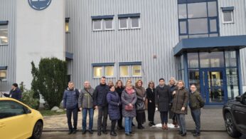 Grupa 15 osób, ubranych w zimowe ubrania, stoi i pozuje razem przed nowoczesnym szarym budynkiem przemysłowym z niebieskimi wykończeniami i dużymi oknami. Dwa zaparkowane samochody, jeden żółty i jeden czarny, są widoczne po obu stronach grupy.