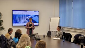 Kobieta w okularach stoi i przemawia do siedzącej grupy w sali konferencyjnej. Na slajdzie za nią widnieje napis 