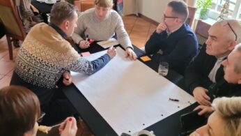 Grupa ludzi siedzi przy stołach w dobrze oświetlonym pomieszczeniu, współpracując nad projektem. Na jednym ze stołów znajduje się duży arkusz papieru, na którym uczestnicy dyskutują i piszą. Widoczne są filiżanki do kawy, talerze i notatniki, a w tle słychać rozmowy innych osób.