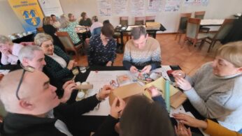 Grupa dorosłych siedzi przy stołach w jasnym pokoju, zaangażowana w praktyczne zajęcia z papierem i markerami. Na ścianie wiszą plakaty z odręcznymi notatkami i diagramami. Ludzie wydają się skupieni i współpracują, pracując razem w warunkach warsztatowych.