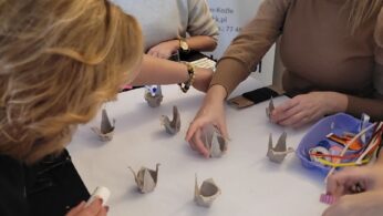 Cztery kobiety siedzą przy stole i składają papier w żurawie origami. Na stole rozrzucone są materiały rzemieślnicze, takie jak kolorowy papier, klej i nożyczki. W tle widoczna jest niebiesko-żółta tarcza i plakaty informacyjne.