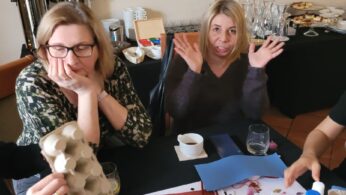 Dwie kobiety siedzą przy stole z przyborami rzemieślniczymi i napojami. Jedna z nich wygląda na znudzoną, opierając głowę na dłoni, podczas gdy druga unosi ręce i uśmiecha się figlarnie. W tle znajdują się tace z jedzeniem, szklanki i dozowniki kawy.