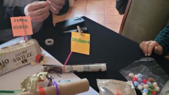 Dwie osoby siedzą przy stole pokrytym materiałami rzemieślniczymi, w tym pomponami, czyścikami do rur i oznaczonymi kartonowymi kreacjami. Widoczna jest odręczna notatka. W tle widać choinkę, roślinę i duże okno z zasłonami.