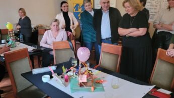 Grupa dorosłych stoi i siedzi wokół stołu pokrytego papierami, markerami i kolorowym modelem wykonanym z balonów, słomek i innych materiałów rzemieślniczych. Wyglądają na zaangażowanych i wesołych w dobrze oświetlonym pokoju z żyrandolem i plakatami na ścianach.