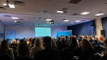 Duża grupa ludzi siedzi naprzeciwko sceny w sali konferencyjnej, obserwując prezentera na podium. Duży ekran wyświetla prezentację z tekstem. Pokój ma niebieskie zasłony, białe ściany, oświetlenie sufitowe i widoczny zegar ścienny.
