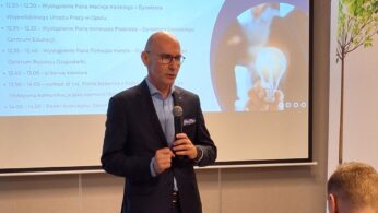 Mężczyzna w garniturze stoi z mikrofonem, przemawiając na konferencji. Za nim ekran wyświetla niebiesko-białą agendę zatytułowaną PROGRAM KONFERENCJI i obraz dłoni trzymającej świecącą żarówkę. Siedzący uczestnicy słuchają na pierwszym planie.