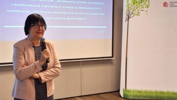 Kobieta z krótkimi ciemnymi włosami, w okularach i wzorzystej kurtce mówi do mikrofonu przed ekranem wyświetlającym slajd prezentacji w języku polskim. Sceneria to sala konferencyjna z białą ścianką działową przedstawiającą ilustrowane drzewo.