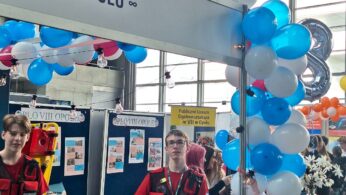 Dwóch nastolatków w czerwonych uniformach stoi przy stoisku wystawowym udekorowanym niebieskimi i białymi balonami. Za nimi widoczne są tablice i plakaty informacyjne. Inni ludzie i więcej balonów wypełniają ruchliwą halę eventową w tle.