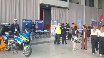 Policyjne stoisko informacyjne z funkcjonariuszami, radiowozem i motocyklem na wystawie. Ludzie, głównie kobiety w swobodnych strojach, gromadzą się w pobliżu stoiska. Widoczny jest żółty znak drogowy z napisem 