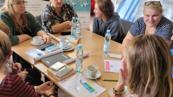 Sześć kobiet siedzi przy stole i dyskutuje, mając przed sobą notatniki, długopisy, papiery i butelki z wodą. Za nimi znajdują się plakaty z tekstem i grafiką. Kobiety wyglądają na zaangażowane i skupione, co sugeruje spotkanie lub warsztaty.