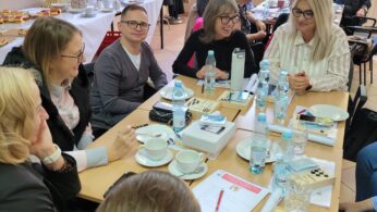 Grupa ludzi siedzi wokół stołu z napojami, przekąskami, papierami i prezentami, uśmiechając się i rozmawiając. Więcej osób siedzi przy stołach w tle. Płaszcze wiszą na wieszakach, a światło słoneczne wpada przez duże okna za nimi.