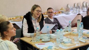 Grupa ludzi siedzi przy stole z notatnikami, butelkowaną wodą, filiżankami i przekąskami. Jedna z kobiet czyta z gazet. W tle znajduje się stół bufetowy z jedzeniem, czajnikami i filiżankami. Atmosfera wydaje się być spotkaniem lub warsztatem.