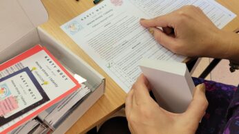 Dwie osoby przy drewnianym biurku korzystające z Kart Mocy dotyczących mocnych stron i talentów. Jedna osoba trzyma talię kart, druga przegląda arkusz roboczy. Widoczne są długopisy, pudełka na karty i materiały drukowane.