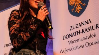 Kobieta o długich brązowych włosach ubrana w czarny koronkowy top i szare dżinsy trzyma mikrofon, stojąc między dwoma banerami. Na prawym banerze widnieje żółty orzeł i napis 