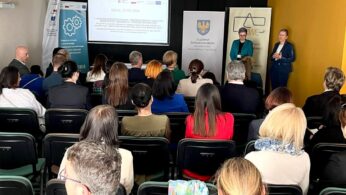 Grupa ludzi siedzi przed ekranem projektora w sali konferencyjnej. Dwie kobiety stoją z przodu, jedna z nich przemawia. Za nimi widoczne są banery i duży slajd prezentacji. Większość uczestników jest odwrócona plecami do kamery, koncentrując się na prezentacji.