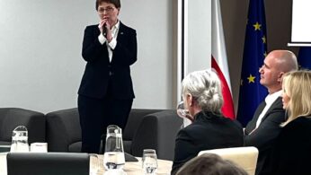 Kobieta w ciemnym garniturze stoi i przemawia przed siedzącą publicznością w sali konferencyjnej. Za nią znajdują się flagi Polski i UE. Czworo słuchaczy, dwie kobiety i dwóch mężczyzn, stoi przed nią. Na pierwszym planie znajduje się stół z dzbankami z wodą i szklankami.