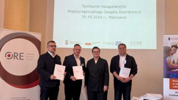 Cztery osoby stoją obok siebie w strojach biznesowych, trzy trzymają certyfikaty. Za nimi ekran projekcyjny wyświetla polski tekst o inauguracyjnym spotkaniu międzyregionalnego zespołu koordynacyjnego w dniu 29.10.2024 r. w Warszawie. Po obu stronach widoczne są dwa zwijane banery.