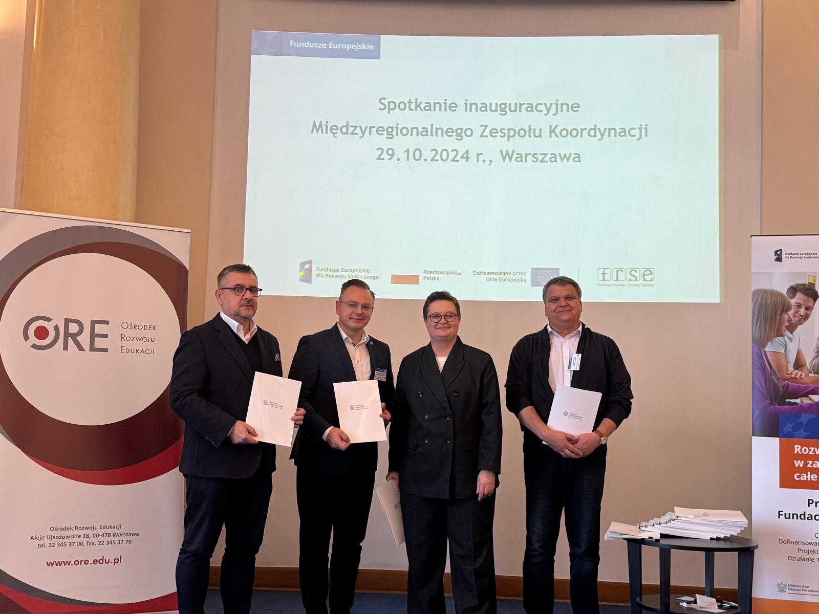 Cztery osoby stoją obok siebie w strojach biznesowych, trzy trzymają certyfikaty. Za nimi ekran projekcyjny wyświetla polski tekst o inauguracyjnym spotkaniu międzyregionalnego zespołu koordynacyjnego w dniu 29.10.2024 r. w Warszawie. Po obu stronach widoczne są dwa zwijane banery.