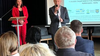 Mężczyzna w szarym garniturze przemawia do mikrofonu przed siedzącą publicznością na konferencji. Kobieta w czerwonej marynarce stoi przy podium. Za nimi ekran projekcyjny wyświetla polski tekst na temat edukacji i rozwoju zawodowego.