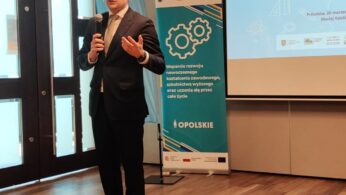 Mężczyzna w garniturze mówi do mikrofonu, stojąc na drewnianej podłodze obok biało-turkusowego baneru informacyjnego. Za nim ekran projektora wyświetla polski tekst i grafikę sprzętu. Naturalne światło wpada przez duże okna po lewej stronie.