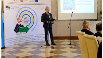 Mężczyzna w garniturze przemawia na podium w sali konferencyjnej, stojąc obok dużego banera z polskim tekstem i logo. Za nim ekran projektora wyświetla slajd. Dwóch siedzących mężczyzn słucha uważnie. Logotypy i informacje o finansowaniu są widoczne u dołu.