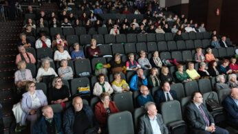 Duża publiczność siedzi w rzędach szarych krzeseł w teatrze lub audytorium, uważnie zwrócona w stronę sceny. Grupa jest zróżnicowana wiekowo, obecni są zarówno starsi, jak i młodsi ludzie. Oświetlenie jest jasne na pierwszym planie i słabsze z tyłu.