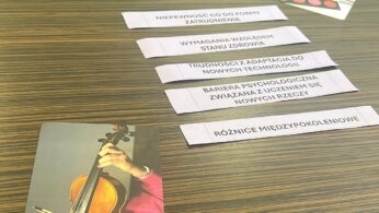 Na drewnianym stole sześć białych pasków papieru z polskim tekstem opisuje wyzwania, takie jak nowe zatrudnienie, zdrowie, technologia i luki pokoleniowe. W pobliżu znajduje się karta ze zdjęciem osoby grającej na skrzypcach, a inna mała karta przedstawia czerwone przyciski w siatce.