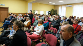 Grupa starszych dorosłych siedzi uważnie w rzędach czerwonych krzeseł w dobrze oświetlonym pokoju, słuchając mówcy. Niektóre osoby noszą okulary i kolorowe ubrania. Jedna osoba z tyłu stoi, robiąc zdjęcie lub nagrywając film telefonem.