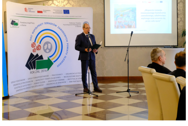 Mężczyzna w garniturze przemawia na podium w sali konferencyjnej, stojąc obok dużego banera z polskim tekstem i logo. Za nim ekran projektora wyświetla slajd. Dwóch siedzących mężczyzn słucha uważnie. Logotypy i informacje o finansowaniu są widoczne u dołu.