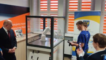 Grupa ludzi obserwuje, jak młody mężczyzna w niebieskiej koszuli obsługuje białe ramię robota wewnątrz szklanej obudowy. Mężczyzna w garniturze i urzędnicy obserwują. W pomieszczeniu znajdują się biało-czerwone rolety okienne w paski oraz banery firmy KUKA i województwa opolskiego.