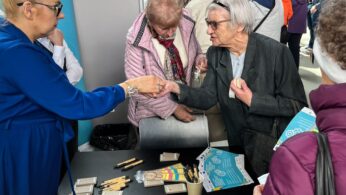 Kobieta w niebieskiej sukience wręcza broszurę starszej kobiecie na zatłoczonym stoisku informacyjnym. Kilku seniorów stoi wokół stołu, na którym leżą ulotki, długopisy i drobne przedmioty. W tle znajduje się więcej osób, tworząc żywą, gwarną atmosferę.