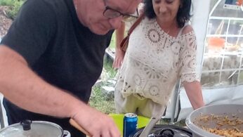 Mężczyzna w okularach i czarnej koszuli podaje jedzenie z dużej patelni, używając drewnianej łyżki. Kobieta w białej koronkowej bluzce stoi w pobliżu, obserwując. Na stole, pod białym namiotem, stoją garnki, puszka coli i miska z jedzeniem.