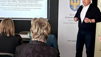 Mężczyzna w ciemnym garniturze przemawia do publiczności w sali lekcyjnej. Za nim znajduje się ekran wyświetlający prezentację w języku polskim z datą 27.03.2024 oraz stojący baner z herbem i napisem 