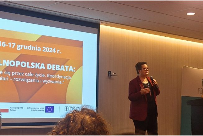 Prelegent stoi z mikrofonem przed ekranem wyświetlającym slajd dotyczący krajowej debaty na temat uczenia się przez całe życie w Polsce w dniach 16-17 grudnia 2024 roku. W sali znajdują się siedzący uczestnicy i baner Fundacji Rozwoju Systemu Edukacji.
