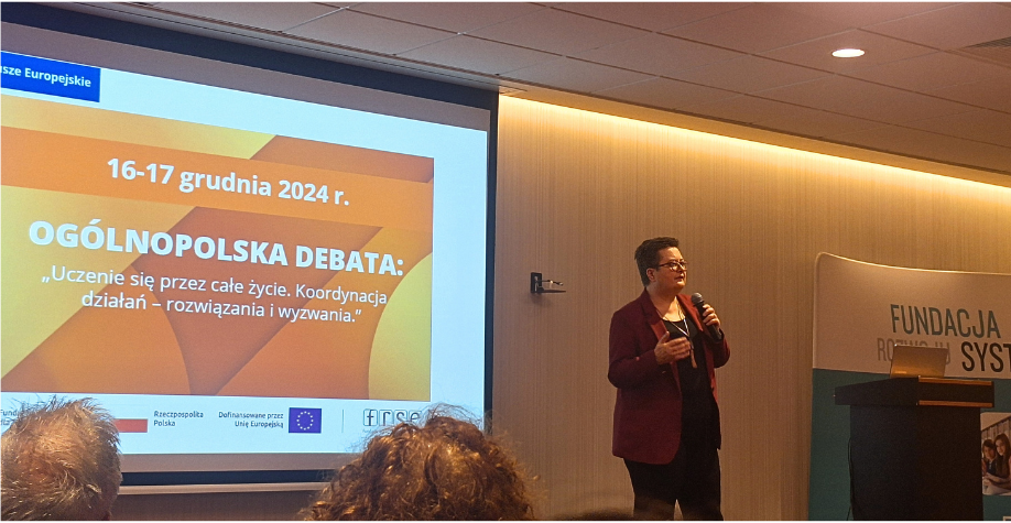 Prelegent stoi z mikrofonem przed ekranem wyświetlającym slajd dotyczący krajowej debaty na temat uczenia się przez całe życie w Polsce w dniach 16-17 grudnia 2024 roku. W sali znajdują się siedzący uczestnicy i baner Fundacji Rozwoju Systemu Edukacji.