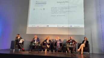 Pięć osób siedzi na scenie podczas konferencji. Po lewej stronie siedzi moderator z mikrofonem i kartką papieru. Czterech panelistów siedzi w fotelach, z mikrofonami, pod ekranem wyświetlającym nazwiska i tytuł konferencji 