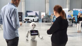 Humanoidalny robot z ekranem wchodzi w interakcję z mężczyzną i kobietą w przestronnym, nowoczesnym salonie z samochodami i stoiskami informacyjnymi w tle. Inne osoby siedzą lub stoją w pobliżu, obserwując scenę.