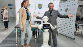 Uśmiechnięty mężczyzna w szarym garniturze i kobieta w brązowej marynarce i białych tenisówkach pozują z białym humanoidalnym robotem na imprezie wewnątrz budynku. Za nimi znajdują się banery informacyjne z logo i polskim tekstem. Inni ludzie chodzą lub stoją w tle.