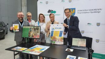 Pięć osób stoi za czarnym stołem, na którym leżą broszury, plakaty i tablety. Dwóch mężczyzn w garniturach i trzy kobiety, wszyscy uśmiechnięci, trzymają materiały promocyjne. Za nimi znajduje się biały baner z polskim tekstem i oficjalnymi logotypami. Sceneria jest wewnątrz budynku.