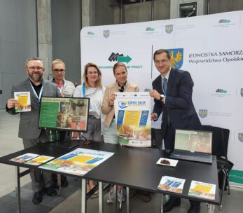 Pięć profesjonalnie ubranych osób stoi za stołem z broszurami, laptopem i plakatami informacyjnymi. Uśmiechają się i prezentują materiały. Za nimi wyświetlany jest duży baner z polskim tekstem i oficjalnymi logo, co sugeruje oficjalne wydarzenie lub targi.