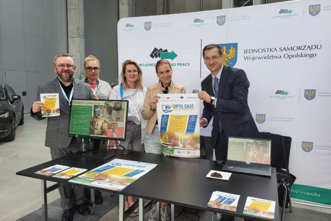Pięć profesjonalnie ubranych osób stoi za stołem z broszurami, laptopem i plakatami informacyjnymi. Uśmiechają się i prezentują materiały. Za nimi wyświetlany jest duży baner z polskim tekstem i oficjalnymi logo, co sugeruje oficjalne wydarzenie lub targi.