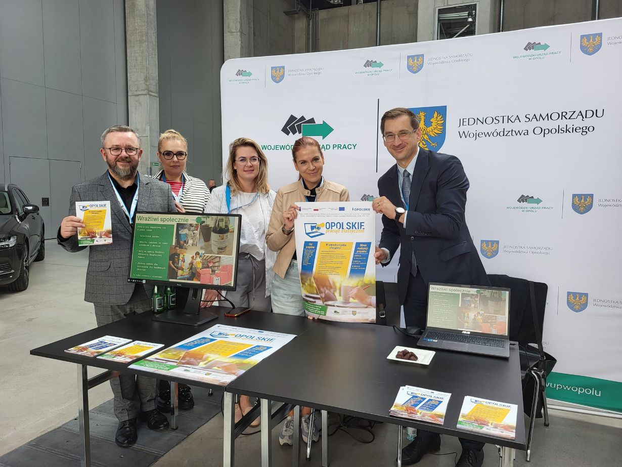 Pięć osób stoi za czarnym stołem, na którym leżą broszury, plakaty i tablety. Dwóch mężczyzn w garniturach i trzy kobiety, wszyscy uśmiechnięci, trzymają materiały promocyjne. Za nimi znajduje się biały baner z polskim tekstem i oficjalnymi logotypami. Sceneria jest wewnątrz budynku.