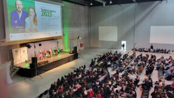 Duże wydarzenie w pomieszczeniu ze sceną, na której cztery osoby siedzą w dyskusji przed publicznością. Na ekranie wyświetlany jest program Týždeň Kaviarny 2023 ze zdjęciami dwóch prezenterów. Rzędy siedzących uczestników wypełniają salę, zwrócone w stronę sceny. Miejsce jest nowoczesne i przestronne.