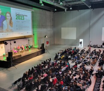 Duże wydarzenie w pomieszczeniu ze sceną, na której cztery osoby siedzą w dyskusji przed publicznością. Na ekranie wyświetlany jest program Týždeň Kaviarny 2023 ze zdjęciami dwóch prezenterów. Rzędy siedzących uczestników wypełniają salę, zwrócone w stronę sceny. Miejsce jest nowoczesne i przestronne.